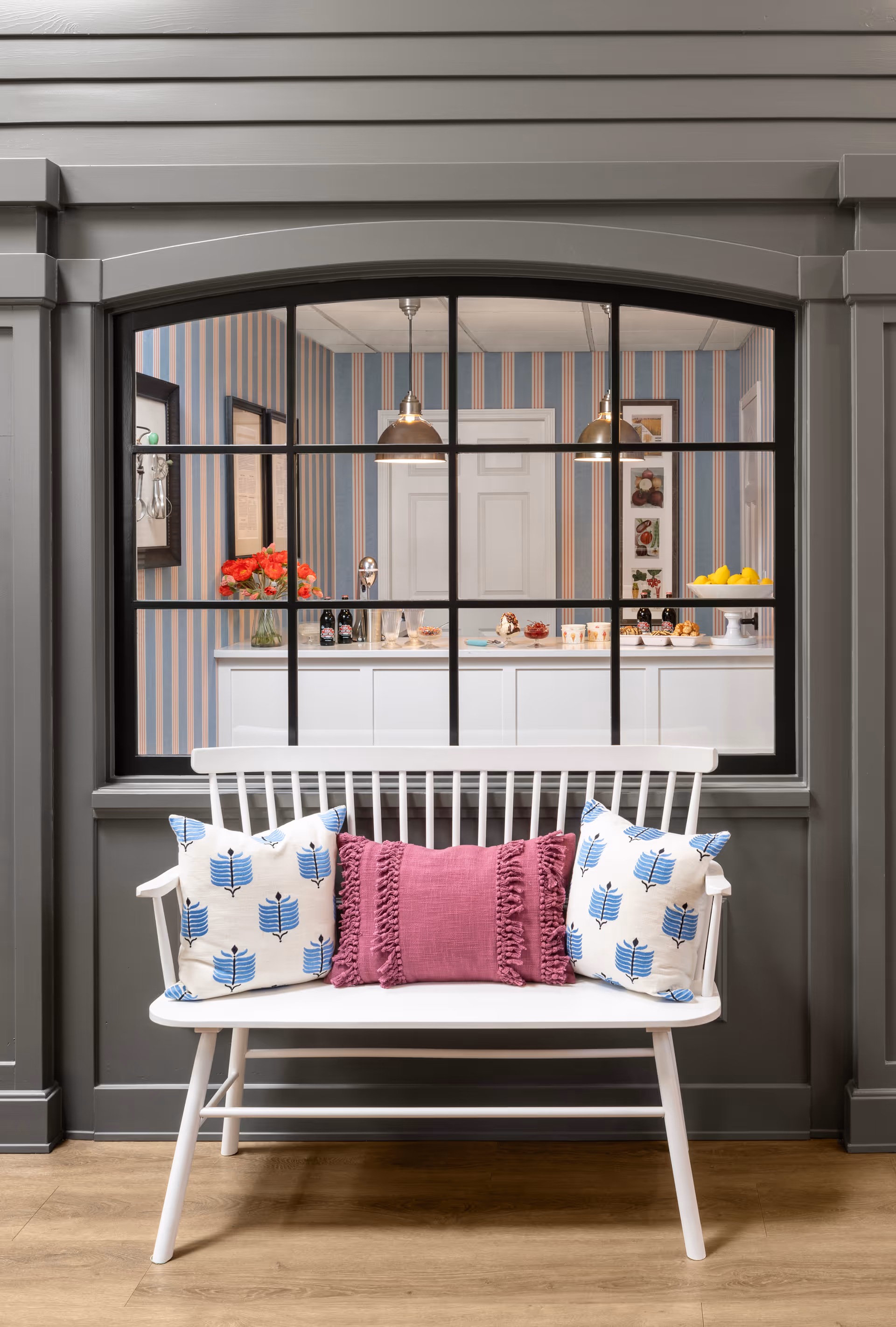 White wooden bench with decorative pillows positioned in front of an interior window looking into a kitchen with pendant lights and striped wallpaper.
