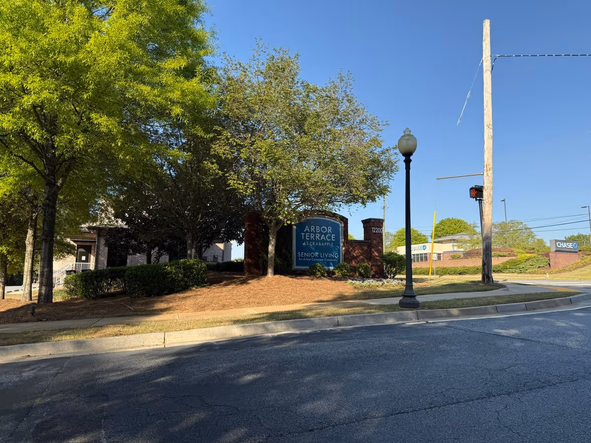 Entrance sign for Arbor Terrace Crabapple senior living surrounded by trees and landscaping beside a street.