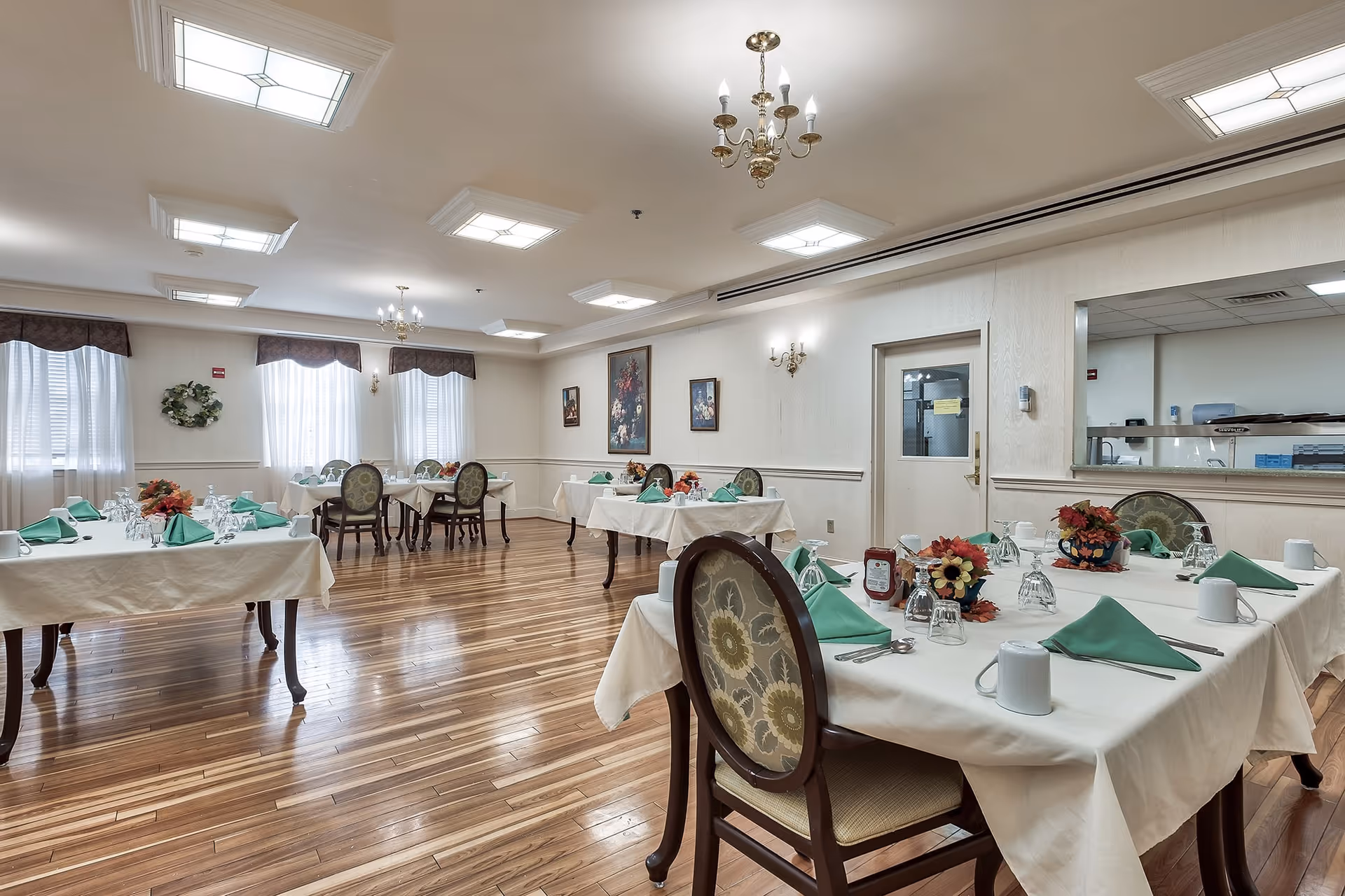 A dining room with several tables covered in white tablecloths, each set with green folded napkins, glassware, white mugs, and floral centerpieces. The room has wooden floors, multiple windows with sheer curtains and valances, chandeliers, and framed artwork on the walls. A door and a large interior window looking into a kitchen area are visible on one side.