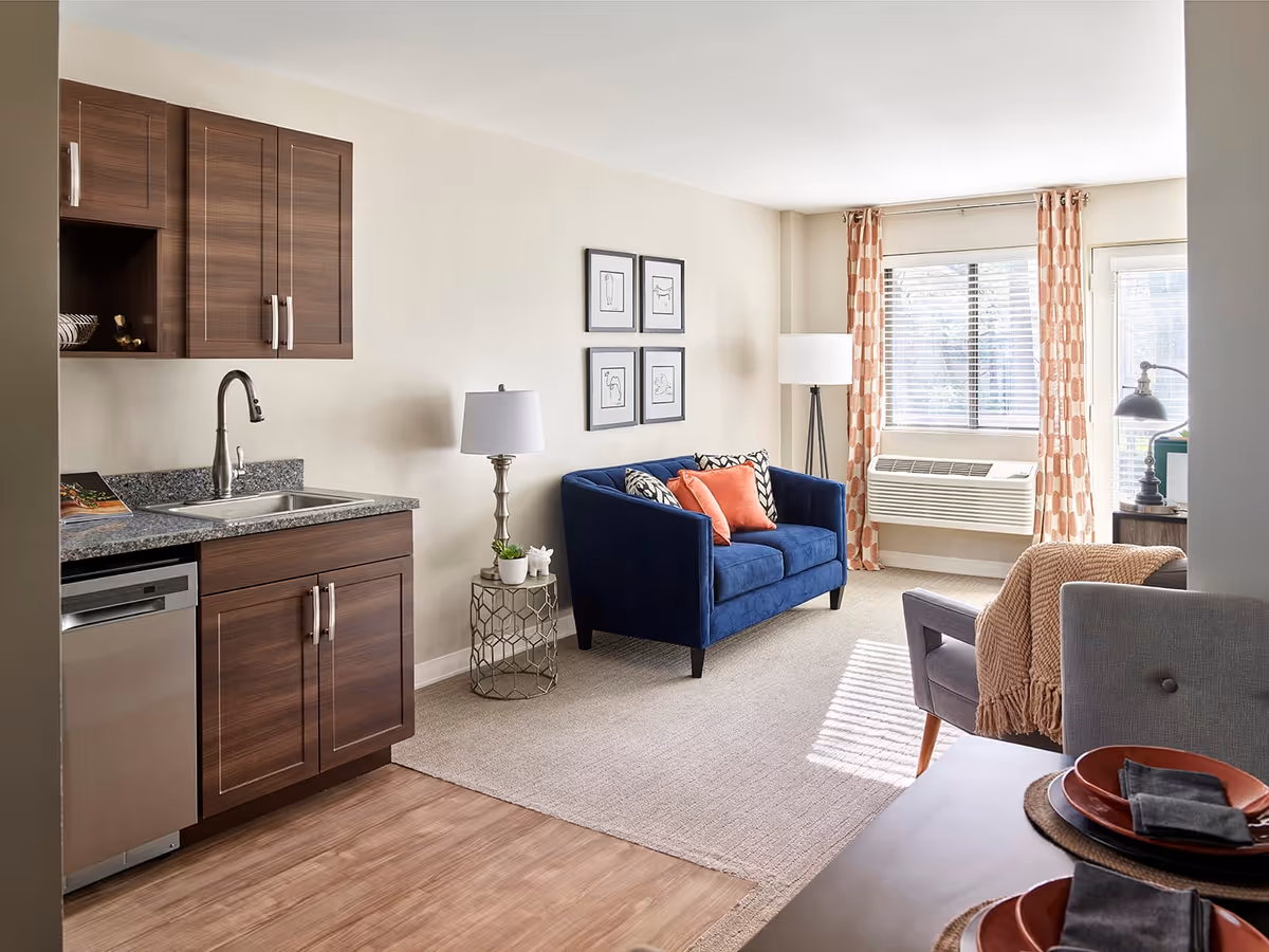 A bright and cozy living area with a small kitchen on the left featuring dark wood cabinets, a sink, and a dishwasher. The living room has a blue loveseat with orange and patterned pillows, a side table with a lamp and small plants, a floor lamp, and large windows with patterned curtains letting in natural light. A dining table with place settings is partially visible in the foreground.