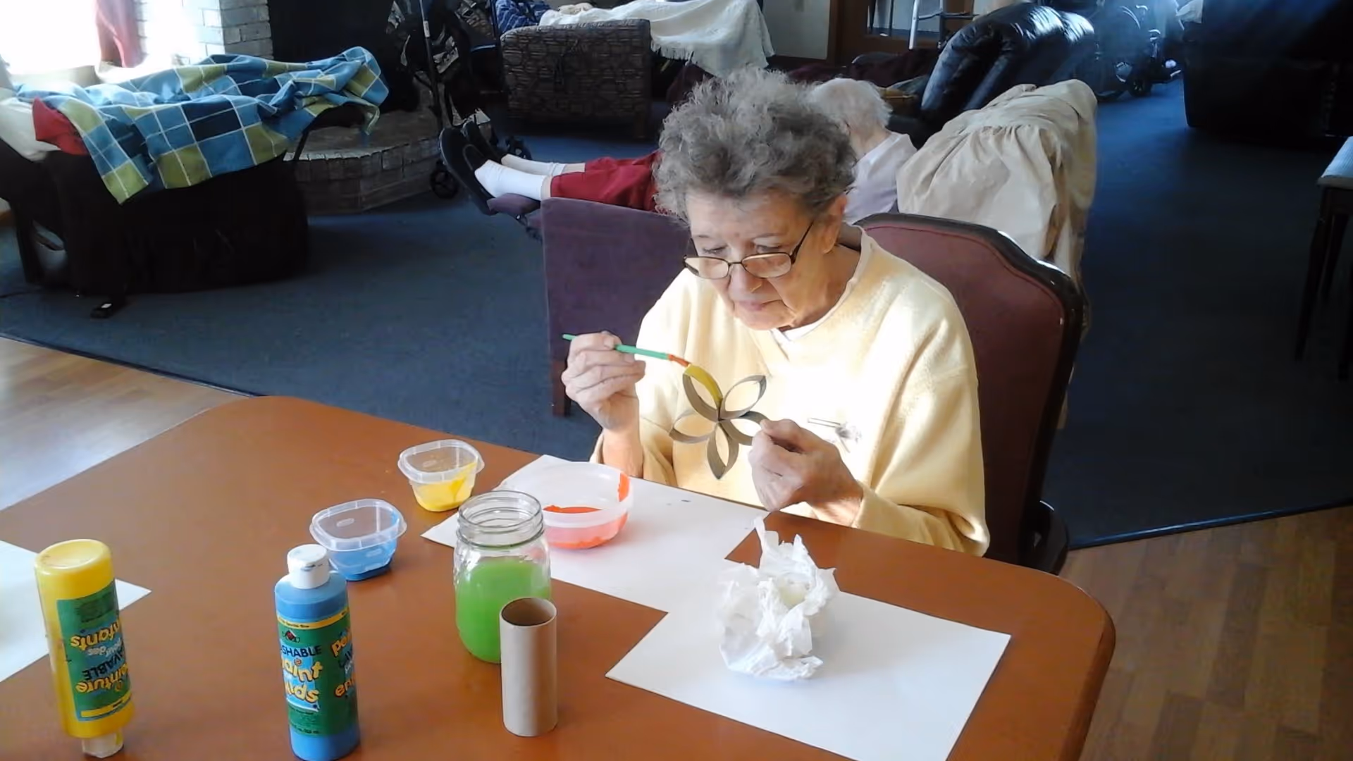 An elderly woman wearing glasses and a yellow sweater is sitting at a table painting a flower-shaped craft. On the table are various painting supplies including bottles of paint, small containers with paint, a jar with green liquid, a cardboard tube, and some paper towels. The background shows a living room area with chairs, a blanket, and other elderly individuals.