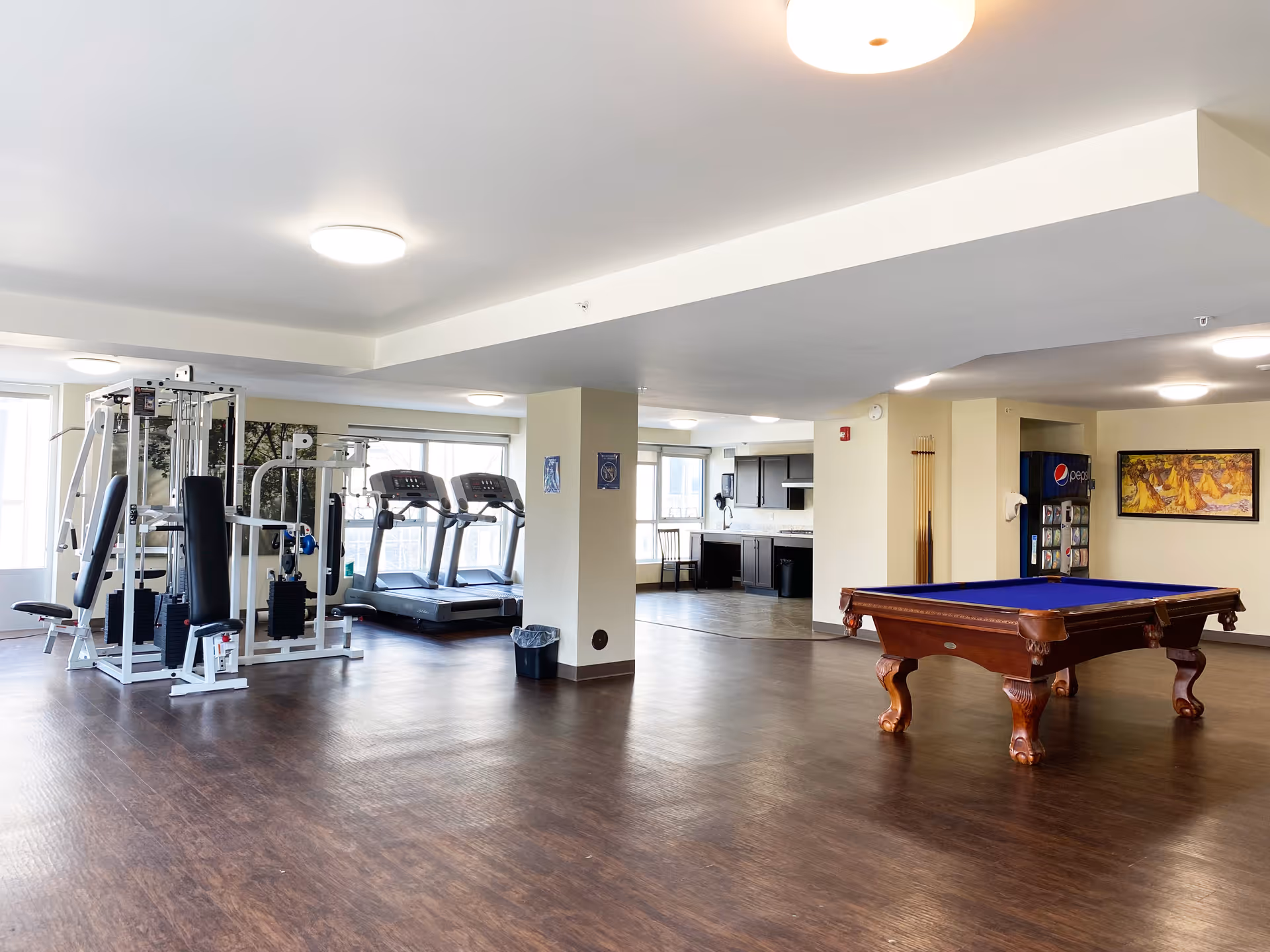 A spacious recreational room featuring exercise equipment including a multi-gym machine and two treadmills near large windows. In the foreground, there is a pool table with a blue felt top. The room has wood flooring, bright ceiling lights, a vending machine, and a kitchenette area with dark cabinets and a chair.