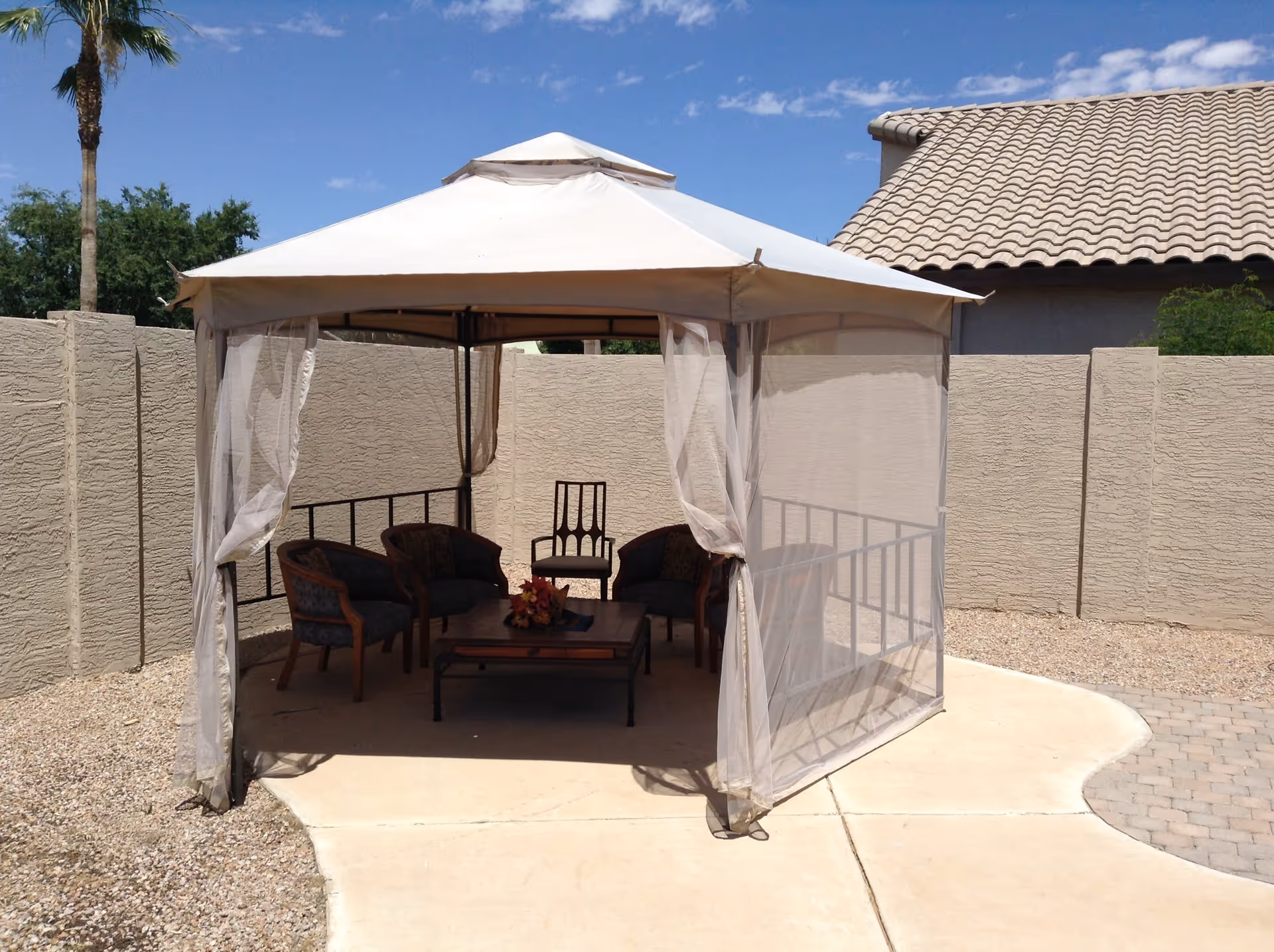 Outdoor gazebo with a white canopy and mesh curtains, containing four cushioned chairs and a wooden coffee table with a flower arrangement, set on a concrete patio surrounded by gravel and a beige stucco wall, with a palm tree and a tiled roof building in the background under a blue sky with scattered clouds.