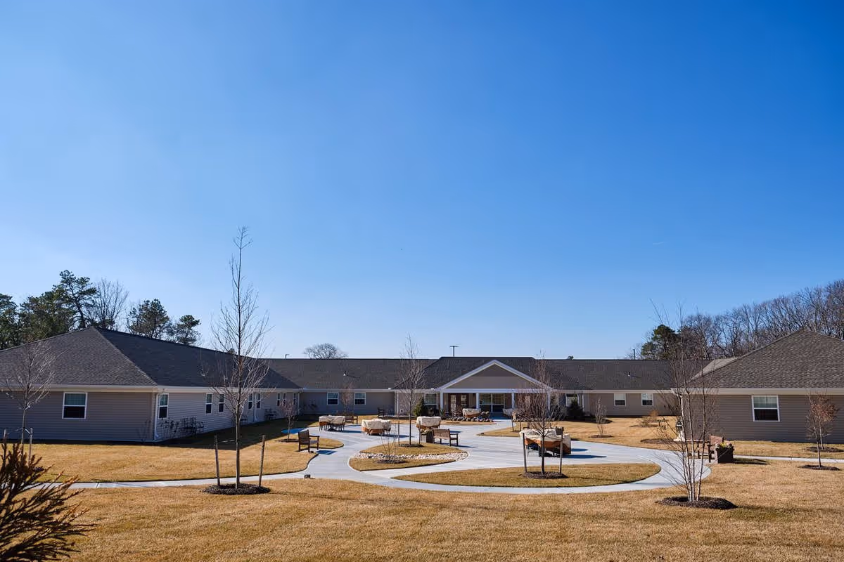 Outdoor courtyard area of a senior living facility with a circular paved walkway, benches, small trees, and a single-story building surrounding the courtyard under a clear blue sky.
