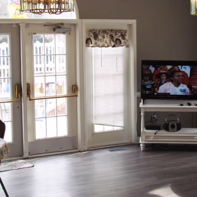 Interior common room with glass double doors to the outside, a tall side window with blinds, and a TV on a white stand.
