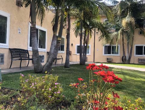 Outdoor courtyard area of a senior living facility with palm trees, green grass, colorful flowers, and benches along the building's beige exterior walls with multiple windows.