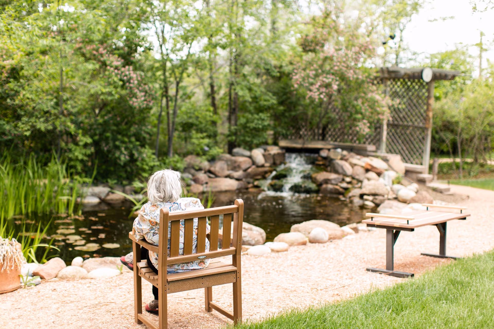 An elderly person with white hair sitting on a wooden bench facing a small pond with a waterfall in a garden surrounded by greenery and trees.