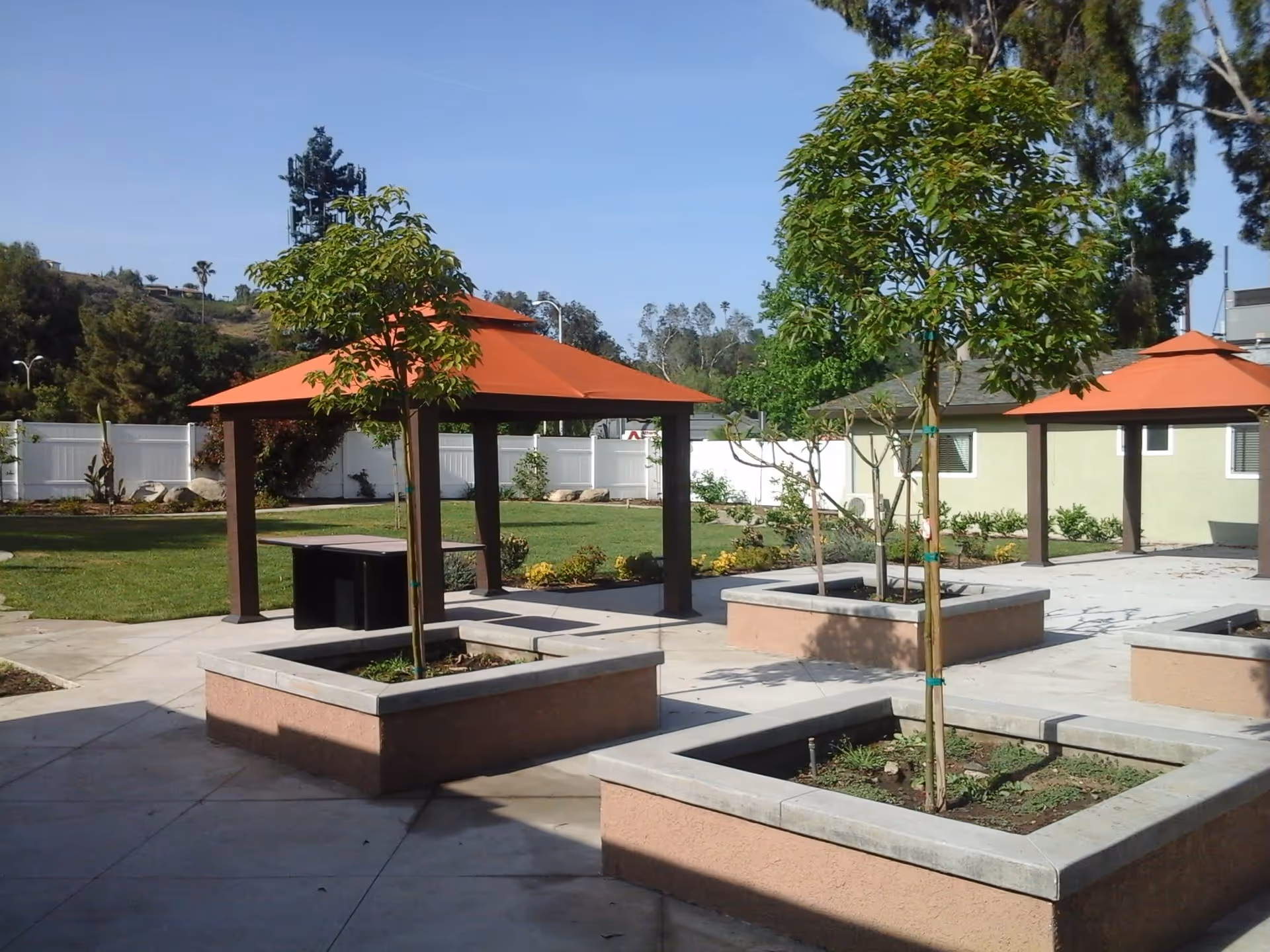 Outdoor area with small trees planted in square raised beds, concrete walkways, and two gazebos with orange roofs. There is a grassy lawn and a white fence in the background under a clear blue sky.