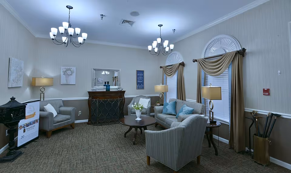 A cozy living room area with beige walls and carpet, featuring two chandeliers, a sofa with blue pillows, three armchairs, two side tables with lamps, a round coffee table with a vase of flowers, two windows with beige curtains, a decorative mirror above a wooden cabinet, and framed artwork on the walls.