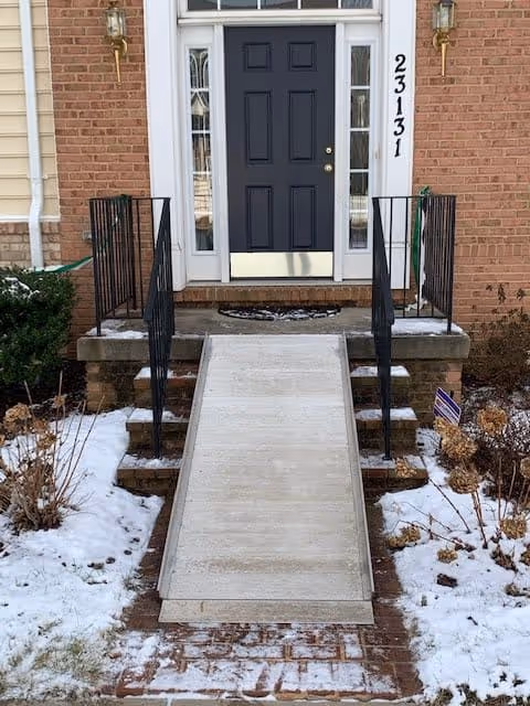 Front entrance of a brick building with a black door, flanked by two narrow windows. There is a concrete wheelchair ramp with black metal railings on both sides, leading up to the door. Snow is visible on the ground and surrounding plants. The building number 23131 is displayed vertically on the right side of the door.