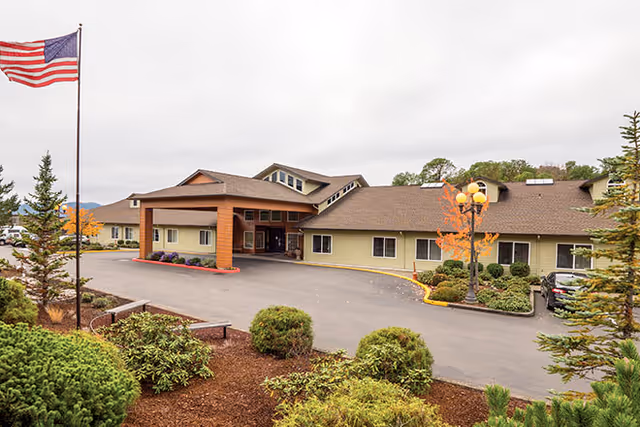 Exterior view of a single-story senior living facility building with a covered entrance, surrounded by landscaped bushes and trees. An American flag is flying on a flagpole to the left, and a street lamp with multiple globes is visible near the entrance. The sky is overcast.