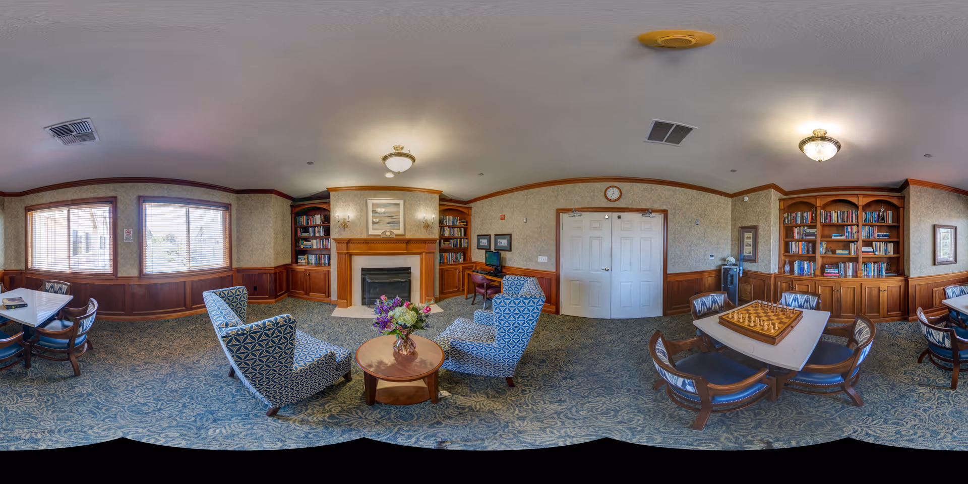 A cozy senior living facility common room with patterned armchairs around a small round table with a flower vase, a fireplace flanked by bookshelves, multiple tables with chairs including one with a chessboard, and windows letting in natural light.