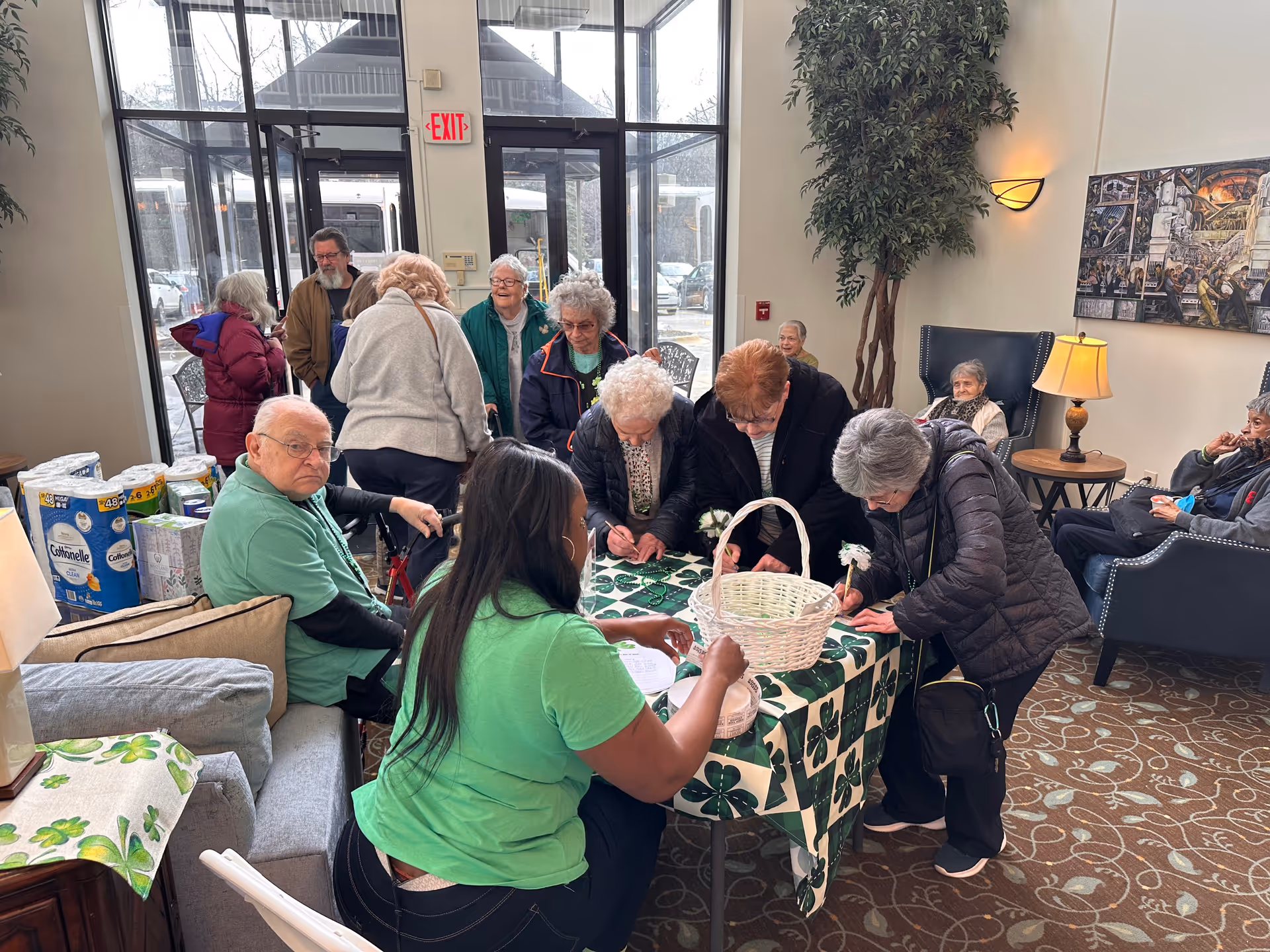 A group of elderly people gathered around a table covered with a green and white clover-patterned tablecloth inside a senior living facility lobby. A woman in a green shirt is seated at the table assisting the elderly individuals who are writing or signing papers. Other seniors are sitting on chairs and couches in the background near large glass doors and windows. The room is decorated with plants, a lamp, and a wall painting.