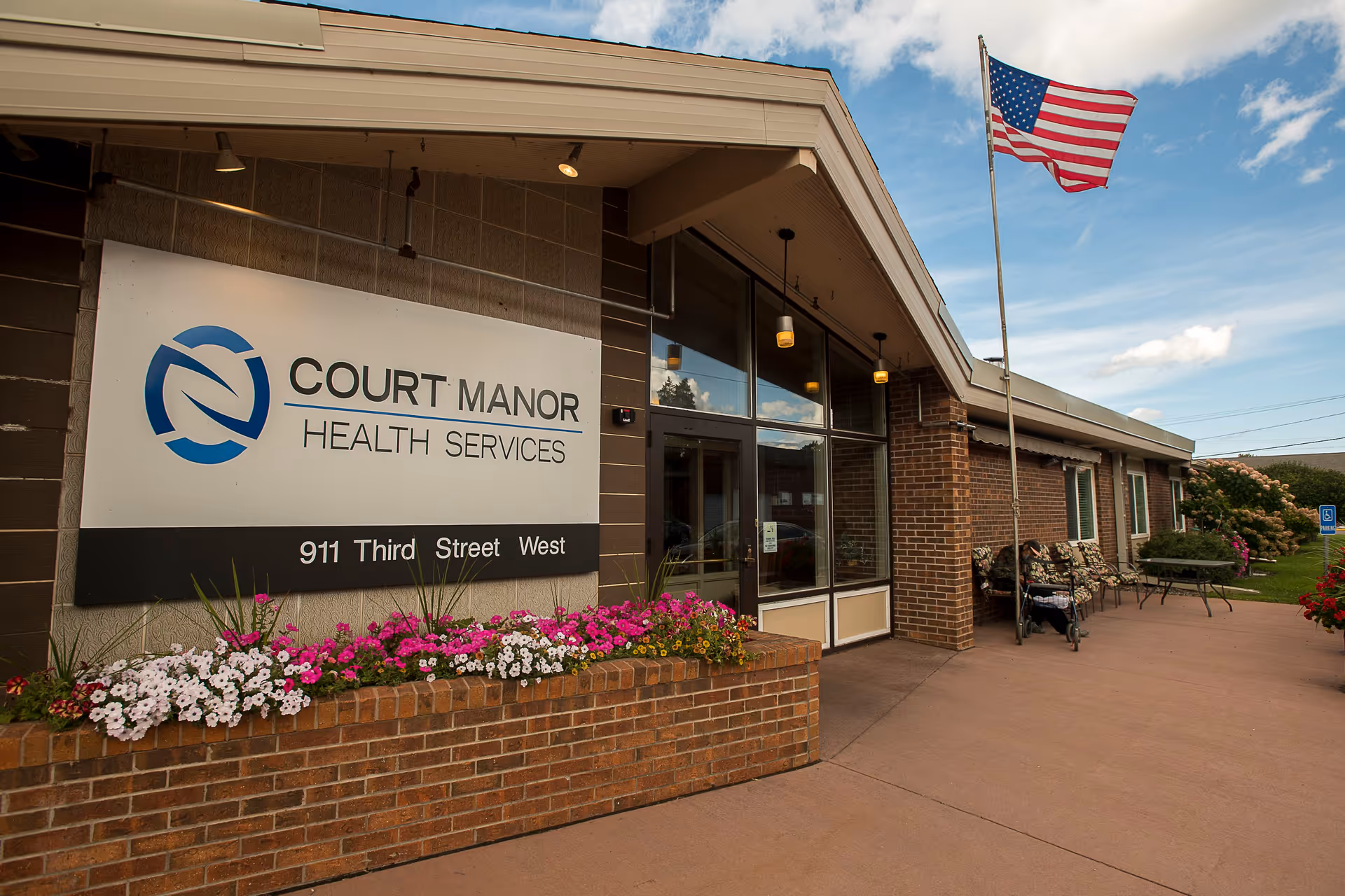 Entrance of Court Manor Health Services with a large sign, flower beds, an American flag, and seating by the doorway.