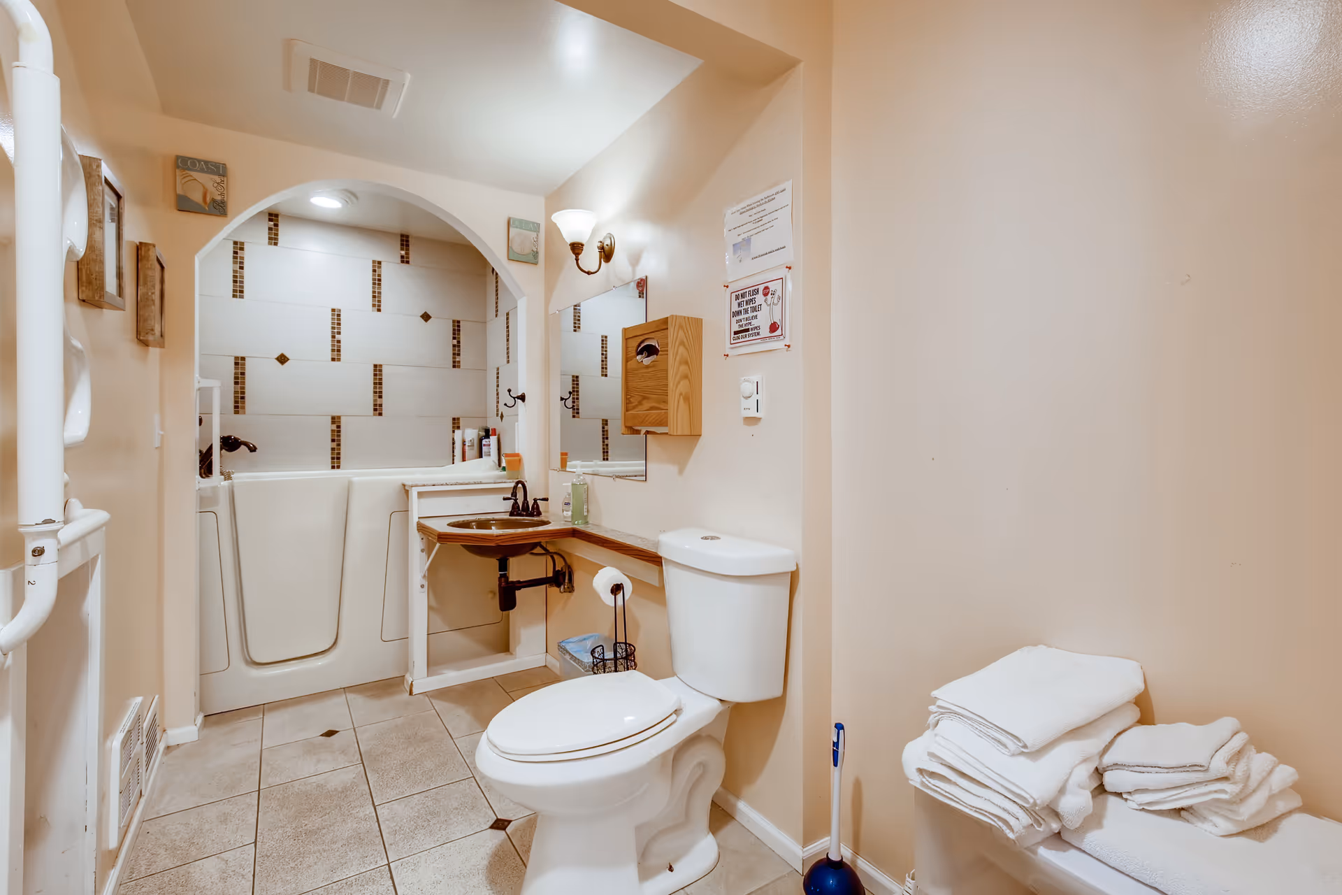 A clean and accessible bathroom featuring a walk-in bathtub with tiled walls, a white toilet, a wooden sink with a mirror above it, a wooden tissue dispenser, and a stack of folded white towels on a bench. The walls are painted beige and the floor is tiled.