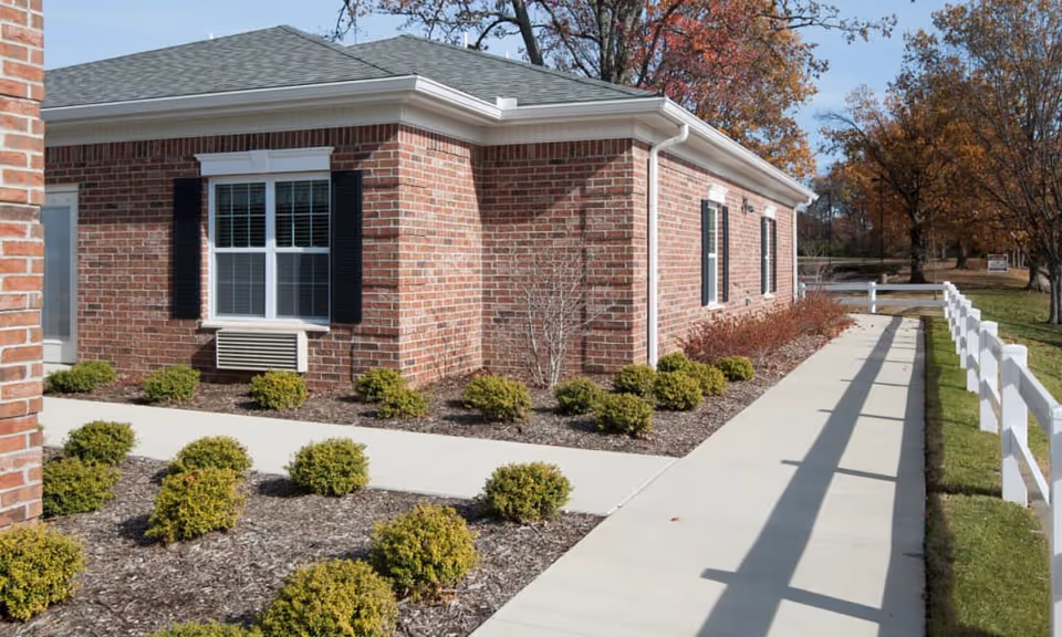 Exterior view of a single-story brick building with white-framed windows and black shutters. There is a concrete walkway bordered by small green shrubs and a white fence on the right side. Trees with autumn foliage are visible in the background under a clear sky.