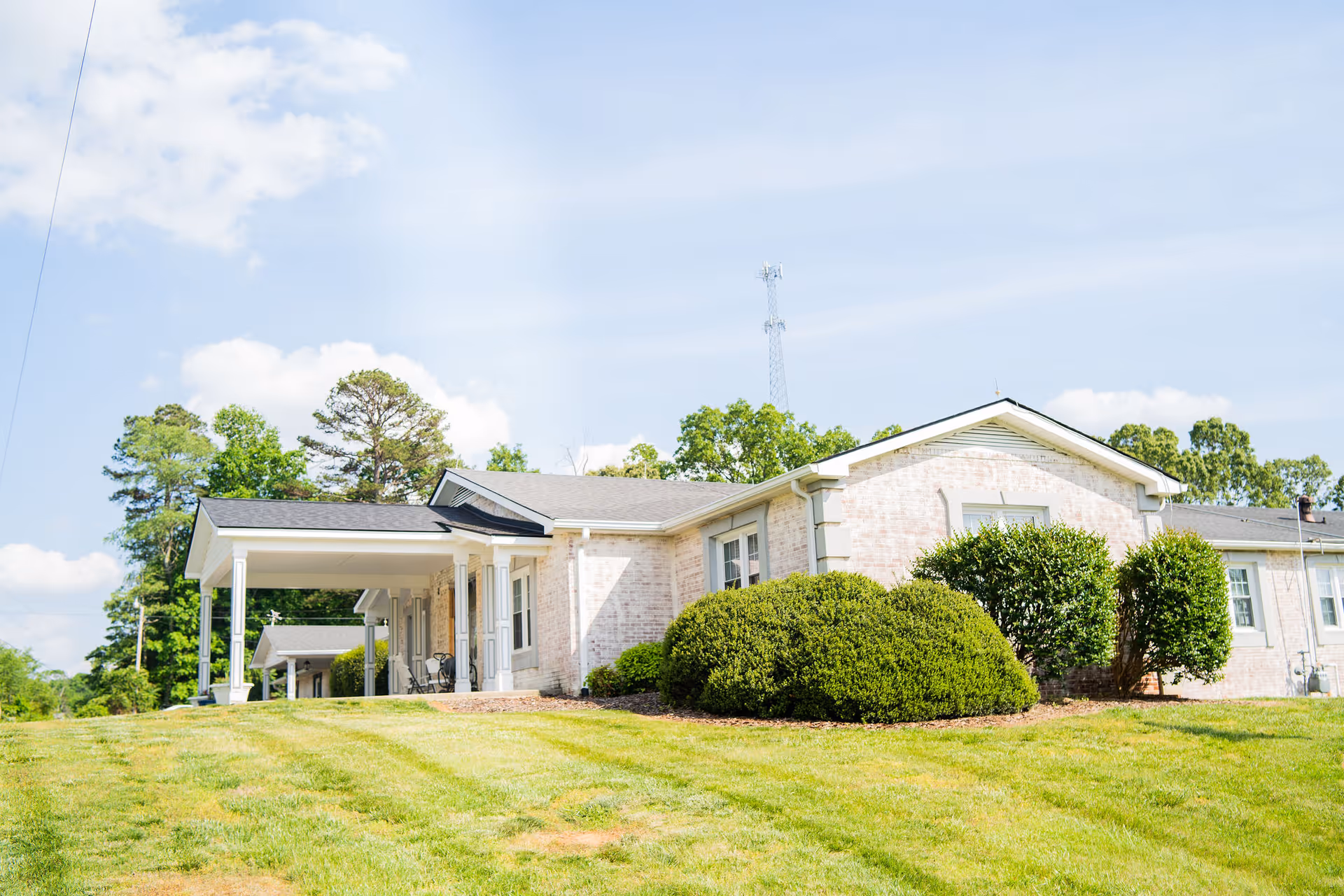 Single-story light-brick memory care building with a covered entrance, shrubs and a mowed lawn under a blue sky.