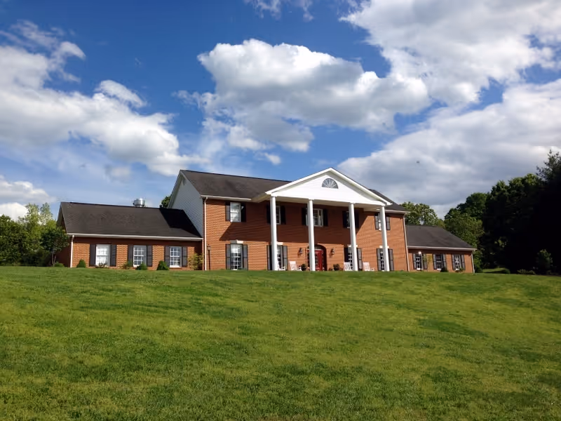 A large two-story brick building with white columns at the entrance, surrounded by a green lawn under a partly cloudy blue sky.