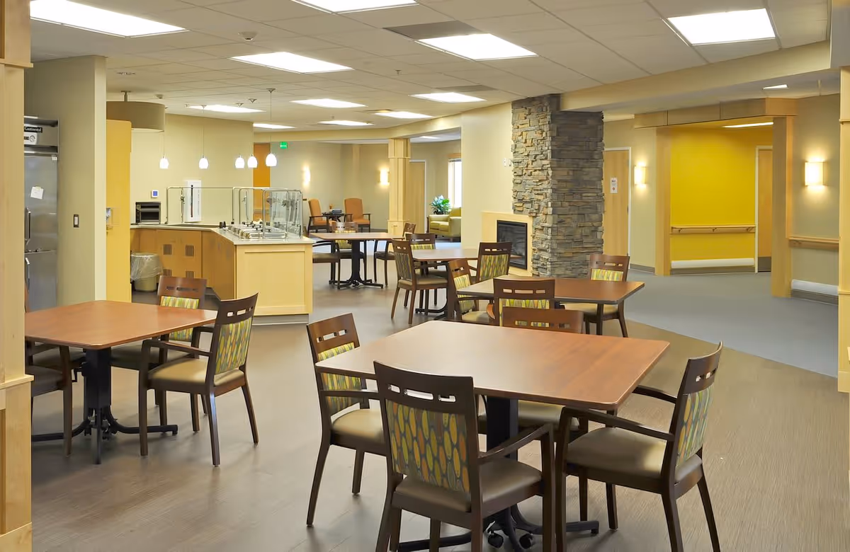 A spacious dining area in a senior living facility with several wooden tables and chairs arranged neatly. The room features a stone fireplace, a serving counter with pendant lights above, and a bright yellow accent wall in the background. The flooring is a combination of wood and carpet, and the ceiling has recessed lighting.