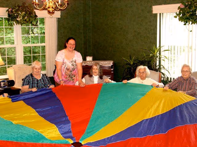 Five elderly women and one younger woman standing and sitting around a colorful parachute indoors in a room with green walls, a window with white trim, and plants. The group appears to be engaged in a recreational activity together.
