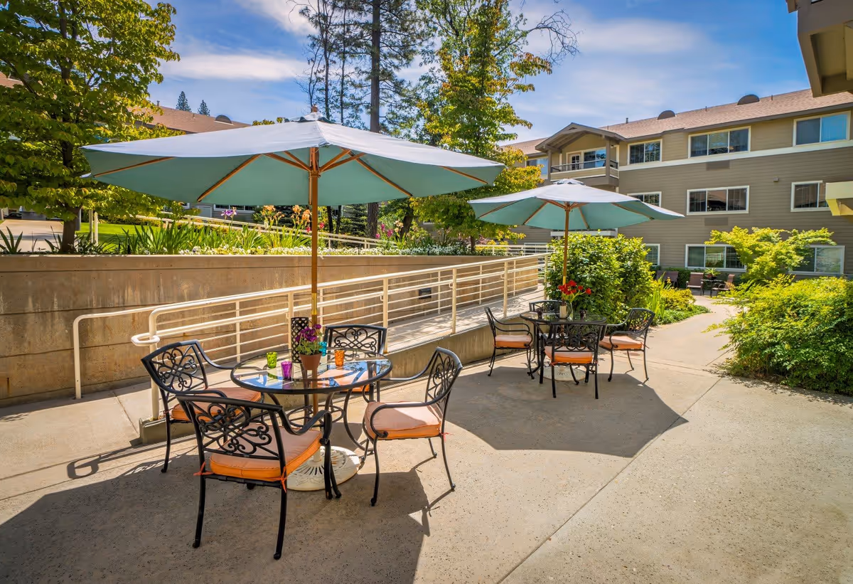 Outdoor patio area at Eskaton Village Grass Valley with two round glass tables, each surrounded by four wrought iron chairs with orange cushions. Each table has a large blue umbrella providing shade. The patio is surrounded by greenery, including bushes and trees, with a beige multi-story building in the background under a partly cloudy blue sky.