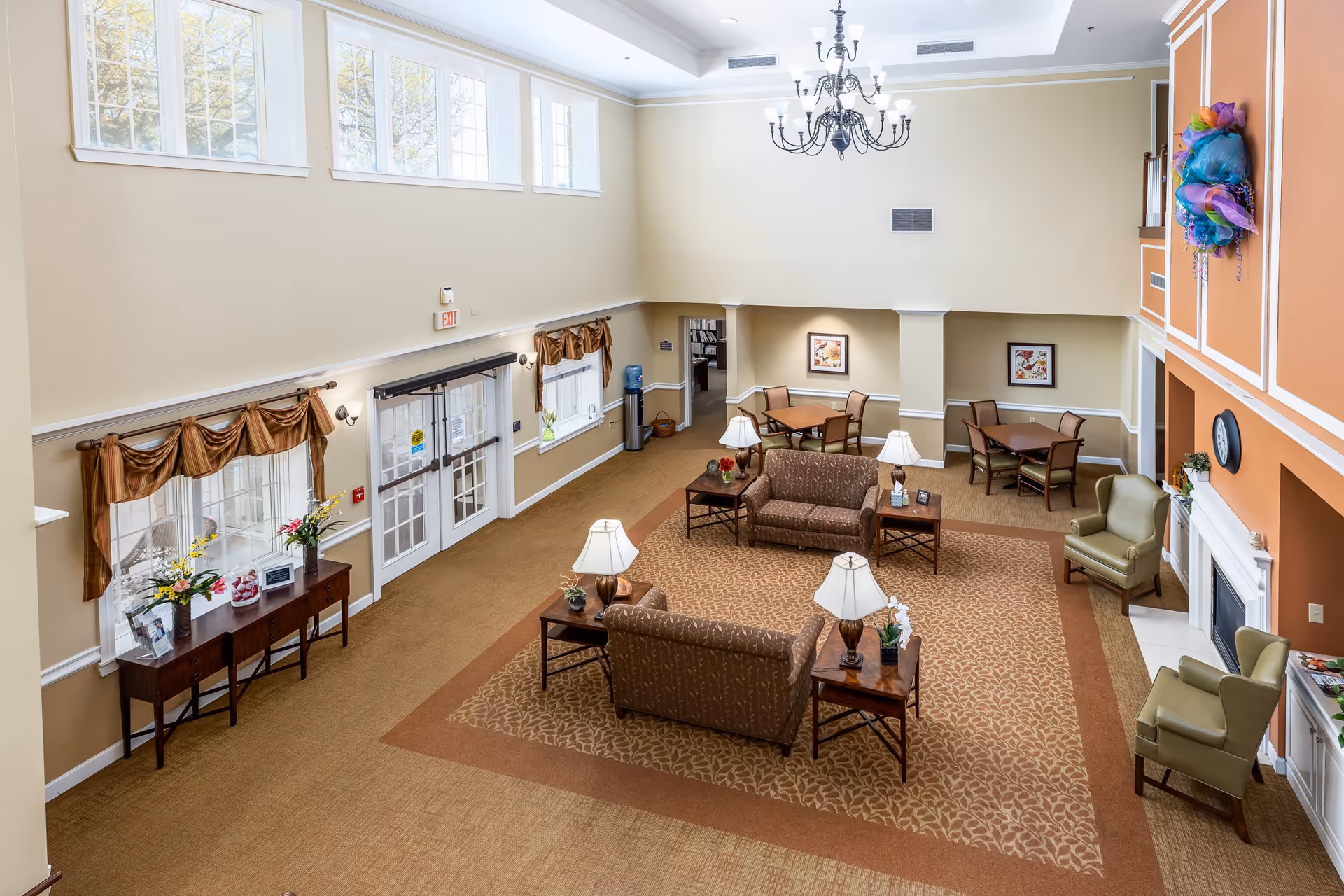 Spacious senior living facility common area with high ceilings, large windows, and a chandelier. The room features two brown patterned sofas, several side tables with lamps, two green armchairs near a white fireplace, and multiple small dining tables with chairs. There is a console table with flowers and framed pictures near the entrance doors, and the walls are painted beige and orange with decorative molding.