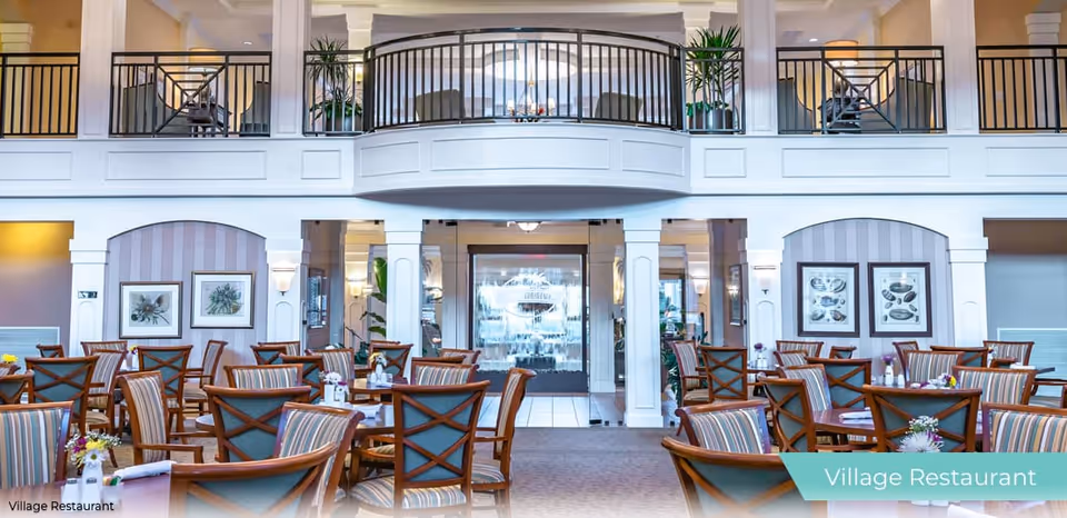 Bright dining room with multiple tables and chairs, an upper balcony, and a central glass water feature.
