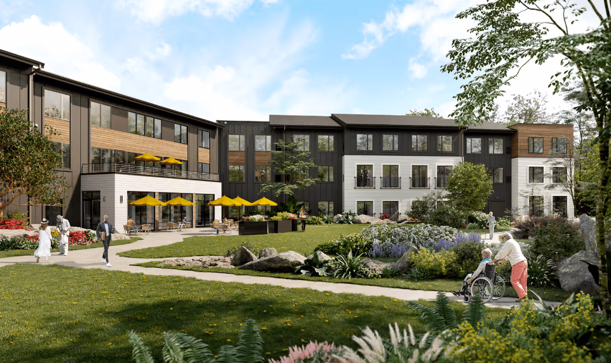 Outdoor view of a senior living facility with a modern three-story building featuring large windows and balconies. The area includes a well-maintained garden with various plants, flowers, and rocks. Several people are visible, including a person pushing another in a wheelchair along a paved pathway, and others walking or sitting under yellow umbrellas on the patio. The sky is partly cloudy with blue patches.