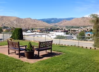 Two wooden benches and a potted plant on a grassy overlook facing a valley with buildings and mountains under a clear blue sky.