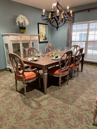 A dining room with a wooden table set for six people, each place setting including a plate, napkin, and glass. The chairs have wooden frames with orange cushions. A white cabinet with glass doors and a flower arrangement on top is against the wall. A chandelier with candle-style lights hangs above the table. A large window with curtains allows natural light into the room.