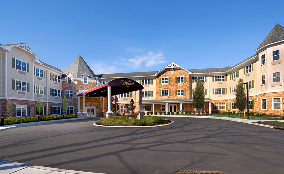 Three-story assisted living building with a covered porte-cochère, central fountain and circular driveway under a clear blue sky.