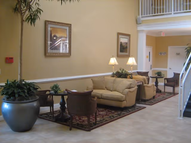 A cozy seating area in a senior living facility with beige sofas, wooden chairs, small tables, table lamps, framed artwork on the walls, and large potted plants. The space has a warm yellow wall color and a tiled floor with area rugs under the seating arrangements.