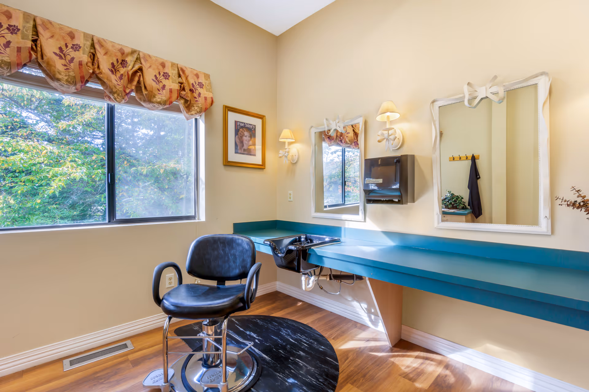 A bright room with a large window showing green trees outside. Inside, there is a black salon chair on a black mat in front of a teal countertop with a built-in black sink. Two white-framed mirrors with decorative bows hang on the wall above the countertop, flanked by two wall-mounted lamps with cream-colored shades. A paper towel dispenser is mounted on the wall between the mirrors, and a framed picture hangs on the adjacent wall.