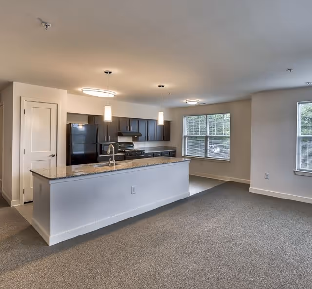 Modern kitchen area in a senior living residence featuring a long granite countertop island with a sink, dark cabinetry, black refrigerator and stove, pendant and ceiling lights, and large windows letting in natural light.
