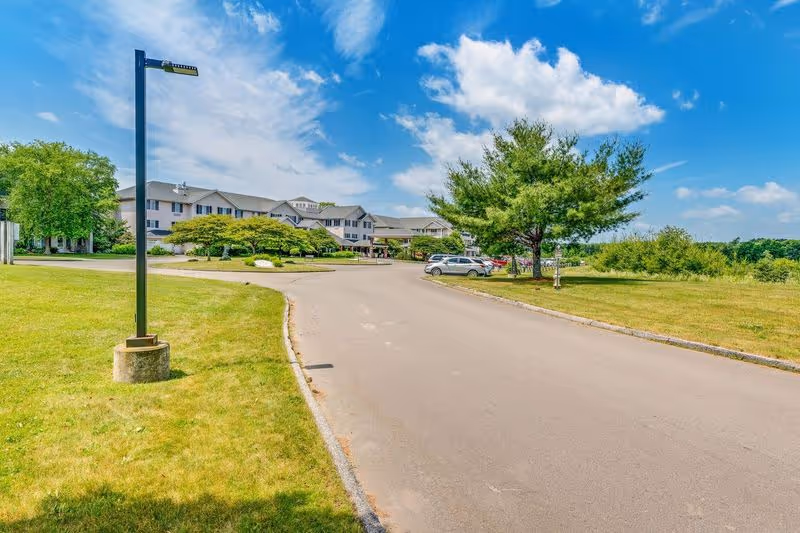 Driveway leading to a multi-story senior living building surrounded by lawns, trees, and parked cars under a blue sky.