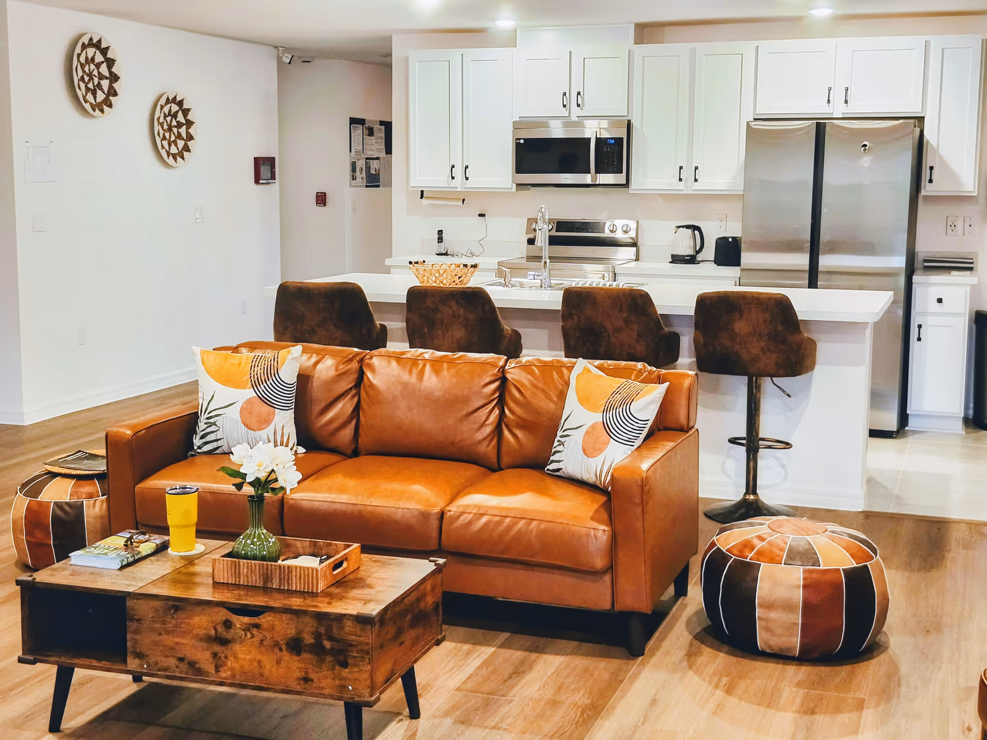 A cozy living room area with a brown leather sofa adorned with two decorative pillows. In front of the sofa is a wooden coffee table with a green vase holding white flowers, a yellow tumbler, and some books. To the left and right of the sofa are two round, multicolored poufs. Behind the sofa is a kitchen with white cabinets, a stainless steel refrigerator, microwave, stove, and a kitchen island with four brown bar stools.
