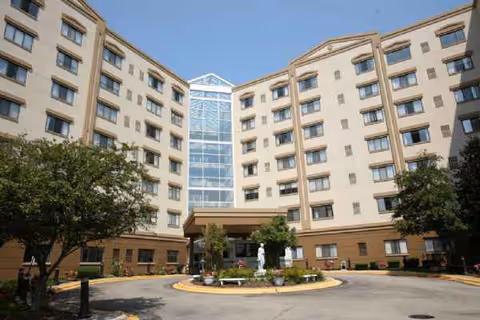 Curved beige senior living building with central glass atrium and circular entry fountain.