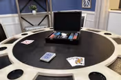 A round black poker table with cup holders and slots for chips. On the table is an open poker chip case with chips and cards, a deck of cards, and a few playing cards spread out. The background shows a blue wall with white trim and a decorative shelf with a small plant.