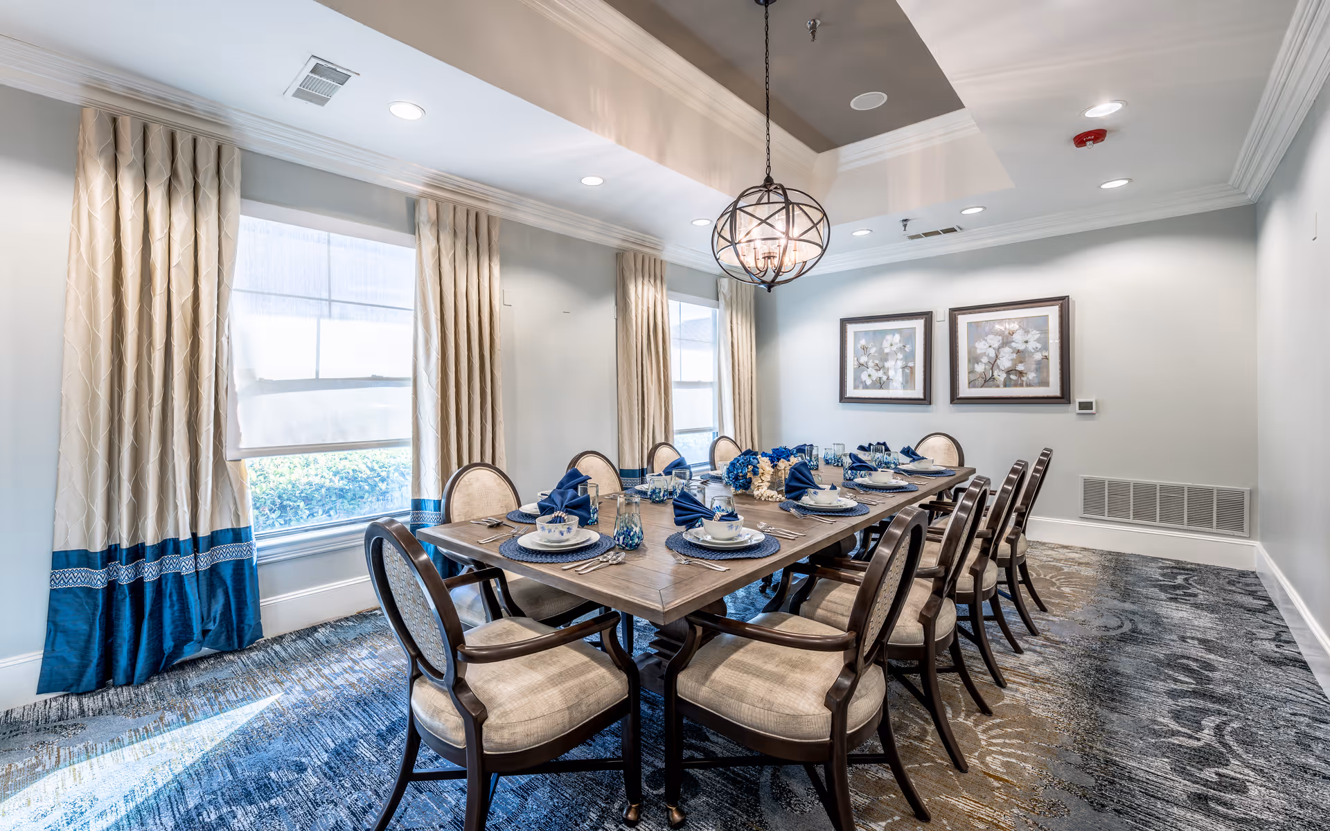 A well-lit dining room with a long wooden table set for twelve people. The table is arranged with blue napkins, white plates, and glassware. The room features large windows with beige and blue curtains, two framed floral artworks on the wall, a patterned carpet, and a decorative hanging light fixture above the table.