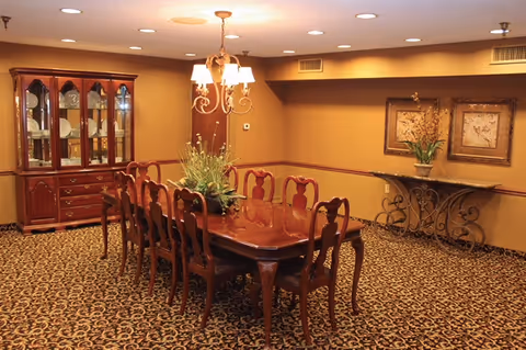 A formal dining room with a long wooden dining table surrounded by eight matching chairs. A chandelier with five lights hangs above the table. There is a wooden china cabinet with glass doors displaying dishes on the left side of the room. On the right wall, there is a decorative console table with a vase of flowers and two framed pictures above it. The room has patterned carpet flooring and warm beige walls.