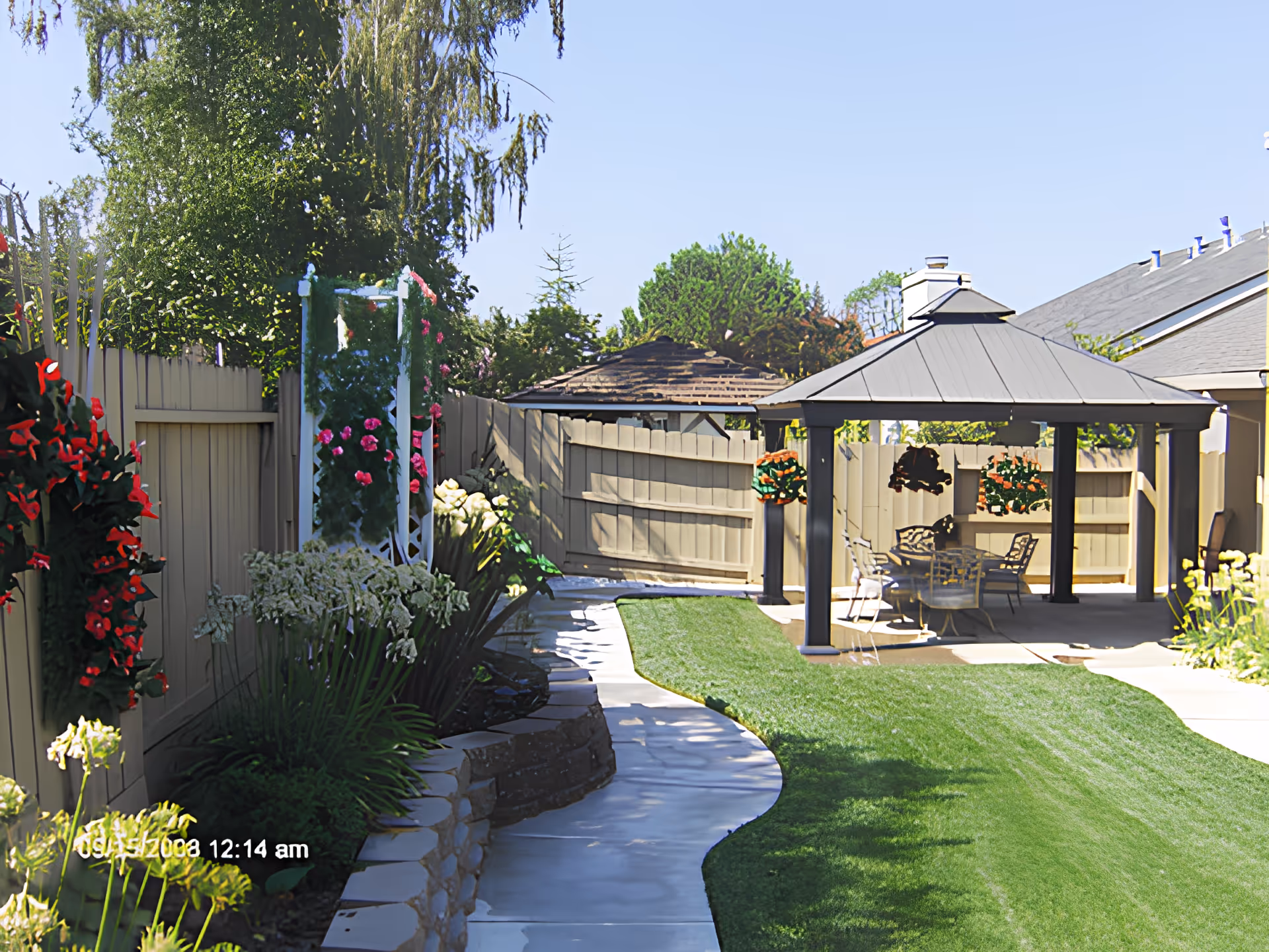 A well-maintained outdoor garden area with a curved stone pathway, green lawn, various flowering plants along a beige fence, and a covered patio with a table and chairs under a gazebo.