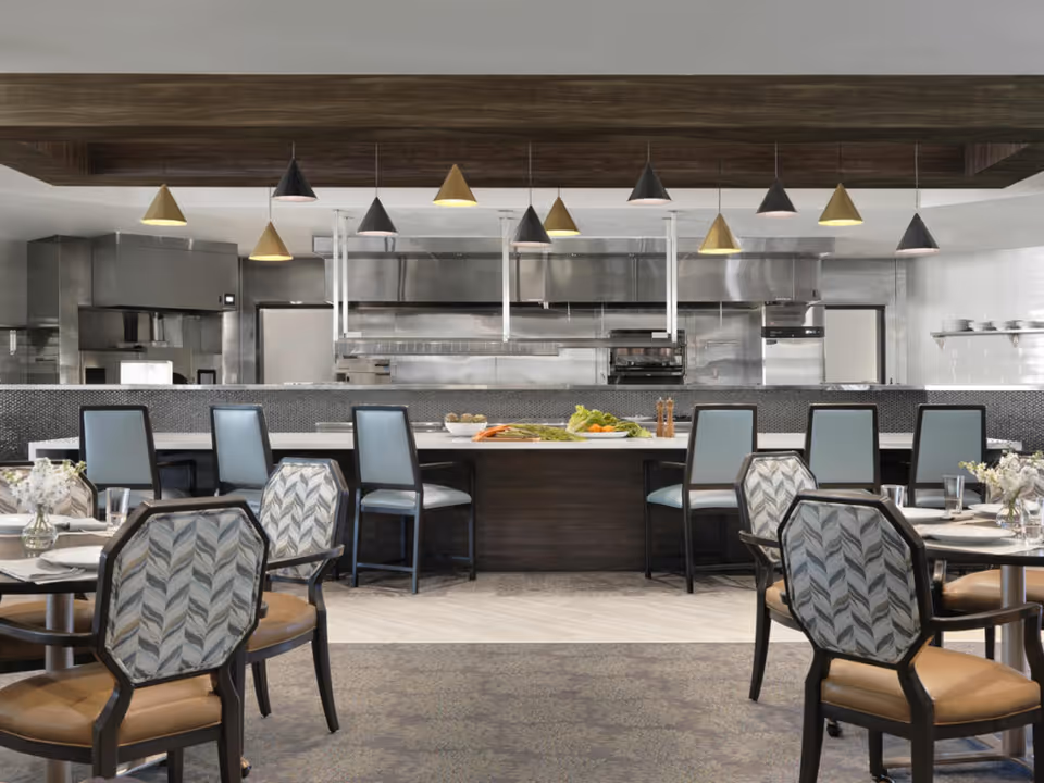 A modern dining area with tables and chairs in the foreground and a large kitchen with stainless steel appliances and a counter with bar stools in the background. Pendant lights hang from the ceiling above the counter.