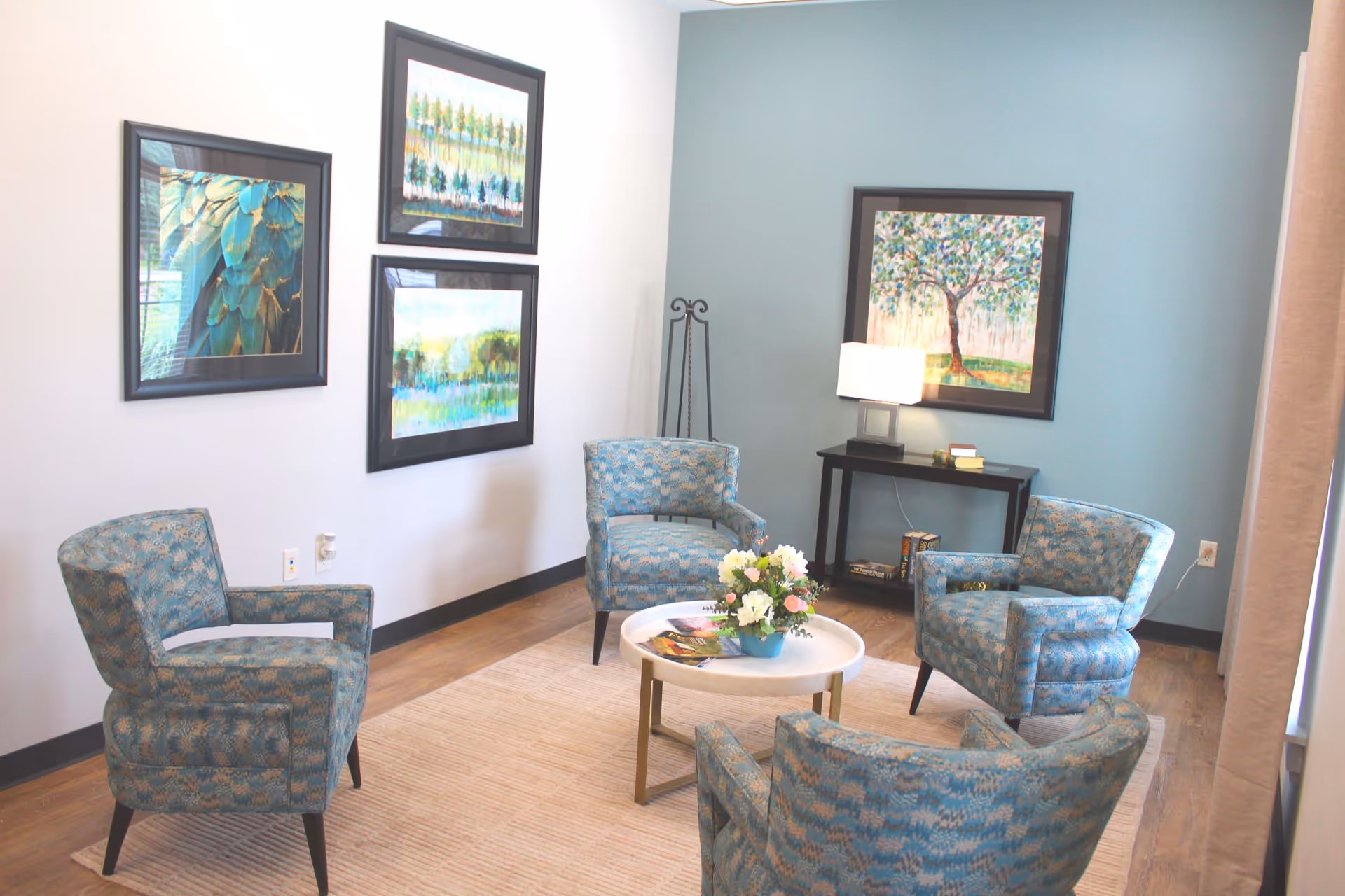 A bright seating area with four patterned armchairs arranged around a round coffee table, artwork on the walls, and a side table with a lamp.
