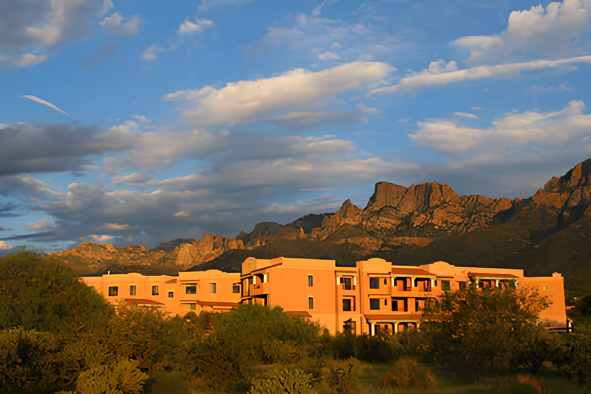 A large, multi-story building with a southwestern architectural style, surrounded by green trees and bushes, set against a backdrop of rugged mountains under a partly cloudy blue sky during sunset.