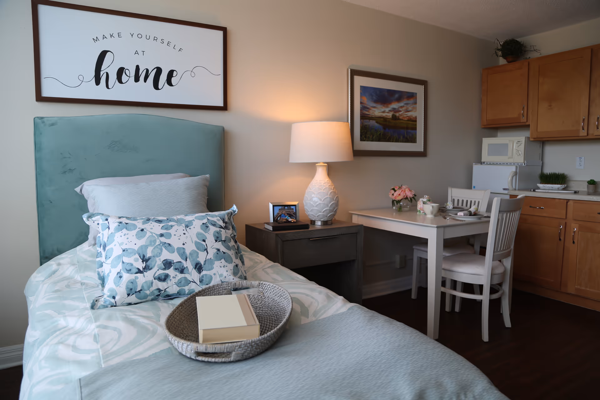 A cozy senior living facility room with a single bed featuring a teal headboard, patterned pillows, and a tray with a book. Next to the bed is a nightstand with a lamp and a framed photo. A small dining table with two chairs is set with flowers and tableware. The room also includes wooden kitchen cabinets, a microwave, and a small refrigerator. A framed picture hangs on the wall, and a sign above the bed reads 'Make yourself at home.'