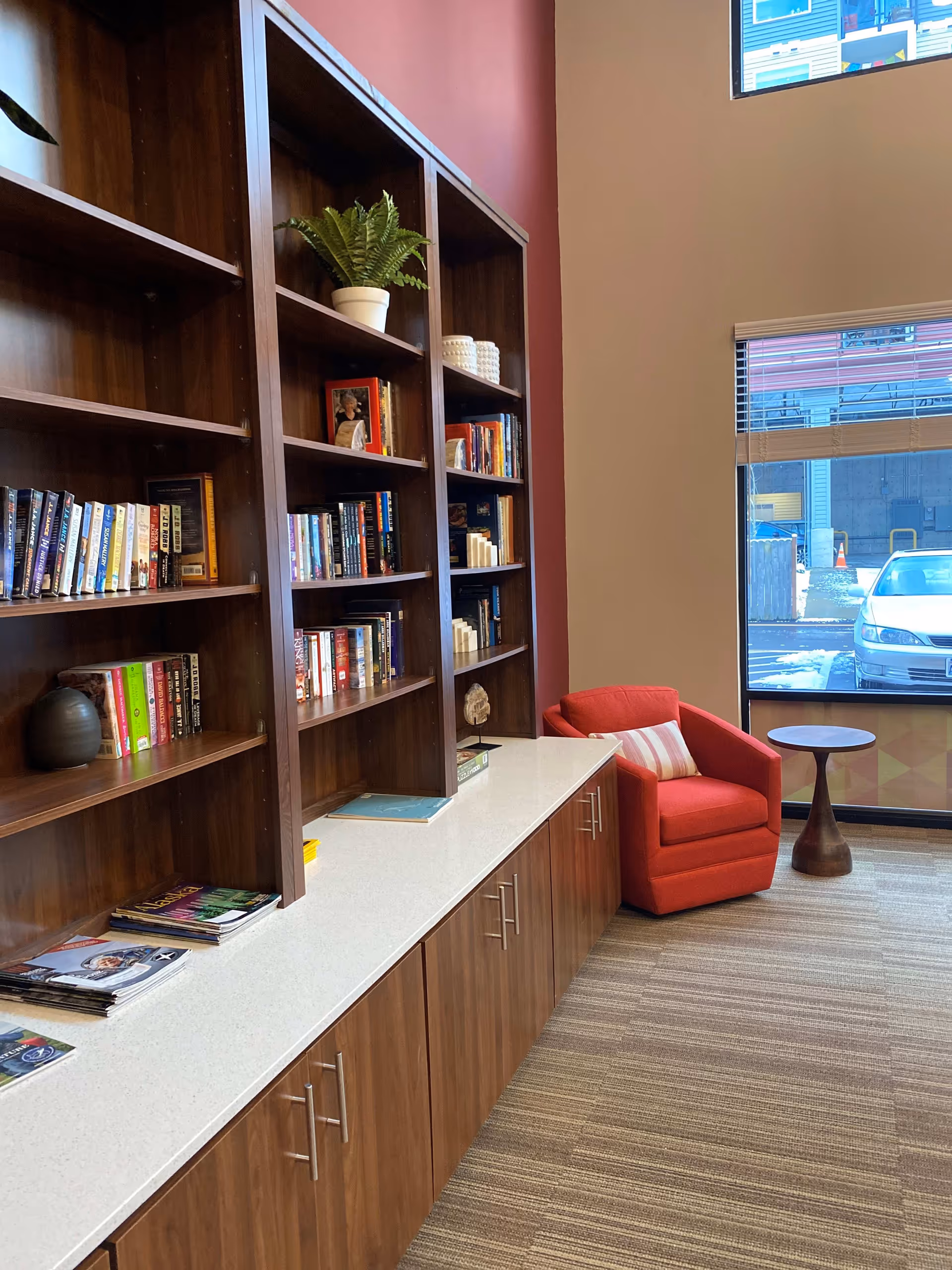 Interior view of a cozy reading nook in a senior living facility featuring tall wooden bookshelves filled with books and decorative items, a red armchair with a striped pillow, a small round wooden side table, and a large window showing a parked car outside.