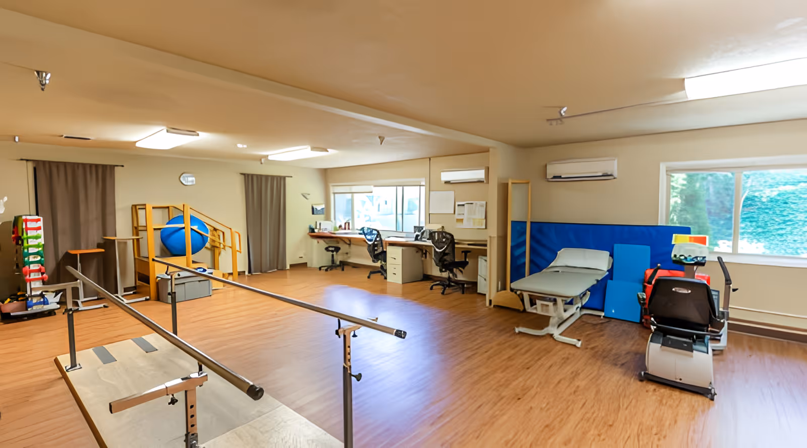 A spacious rehabilitation room with wooden flooring featuring parallel bars for walking exercises, a blue exercise ball, a small set of stairs with handrails, two office desks with chairs near windows, a padded treatment table, and various exercise and therapy equipment.