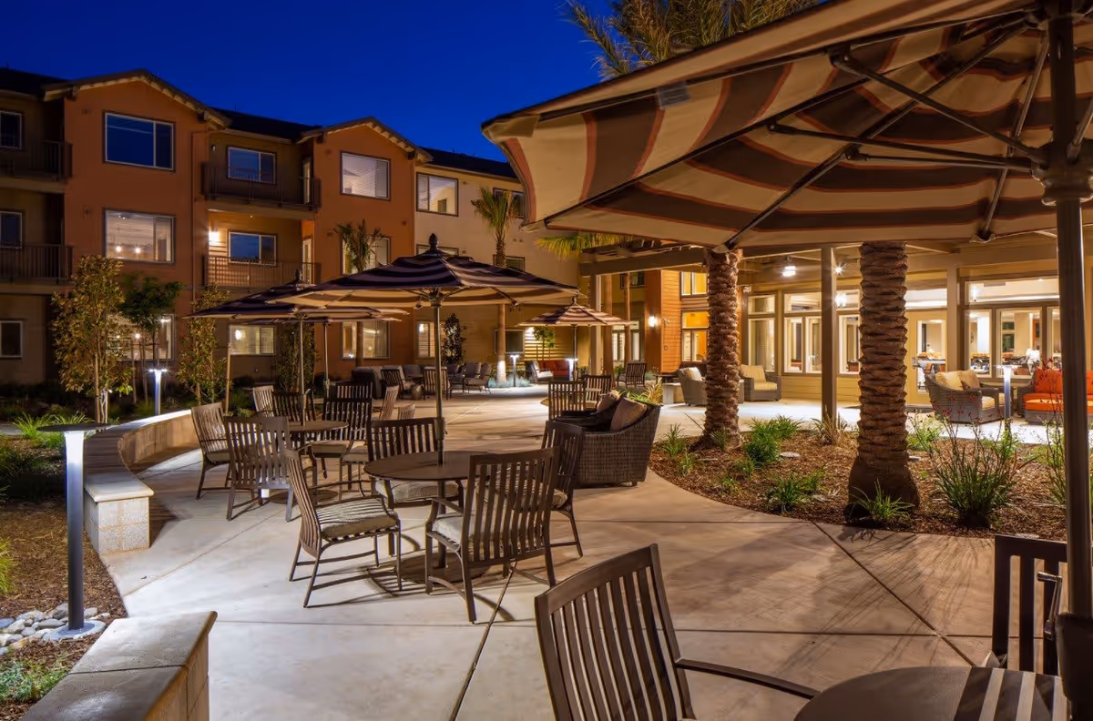 Outdoor patio area at night with multiple tables and chairs under large striped umbrellas. The patio is surrounded by a multi-story building with lit windows and palm trees, creating a warm and inviting atmosphere.
