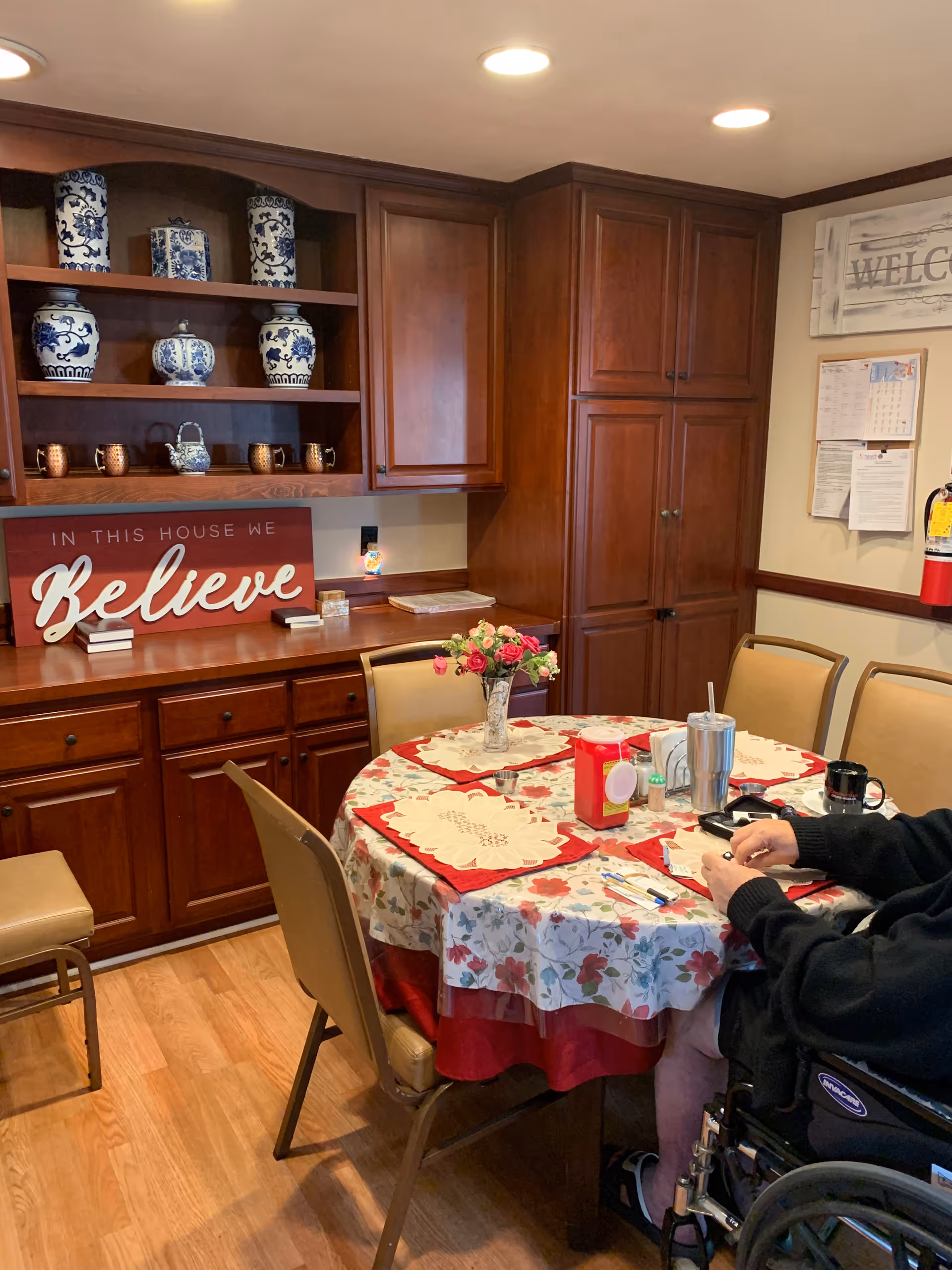 A cozy dining area with a round table covered with a floral tablecloth and red placemats. On the table are a vase with flowers, a tumbler, a coffee mug, and some condiments. Surrounding the table are beige chairs, and a person in a wheelchair is seated at the table. Behind the table is a wooden cabinet with shelves displaying blue and white decorative vases and mugs. A sign on the cabinet reads 'IN THIS HOUSE WE Believe'. The room has warm wooden flooring and recessed ceiling lights.
