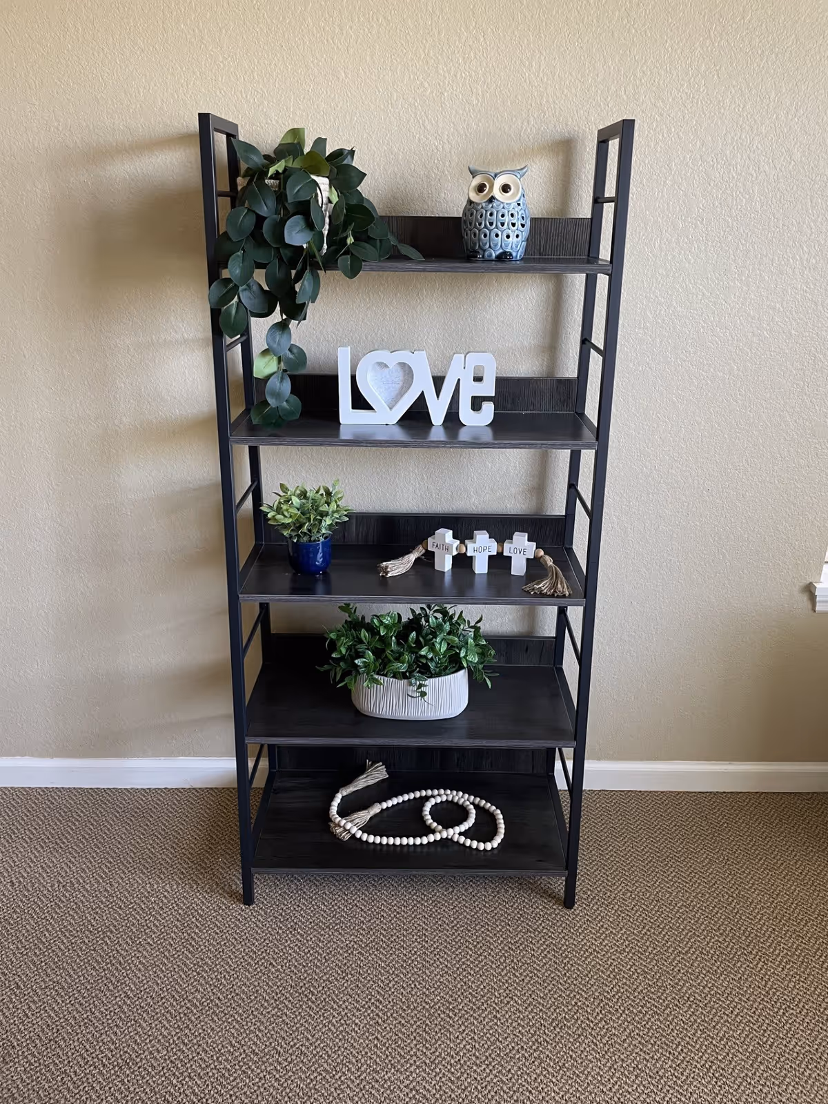 A black metal and wood shelving unit against a beige wall with decorative items including a potted trailing plant, a ceramic owl, a white decorative sign spelling 'LOVE', small potted plants, three small crosses labeled 'FAITH', 'HOPE', and 'LOVE', and a beaded garland on the bottom shelf.